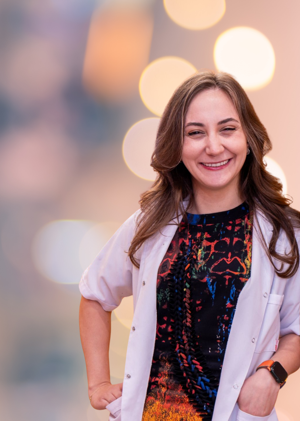 Smiling woman in a white lab coat over a colorful dress, hands in pockets, warm bokeh background.