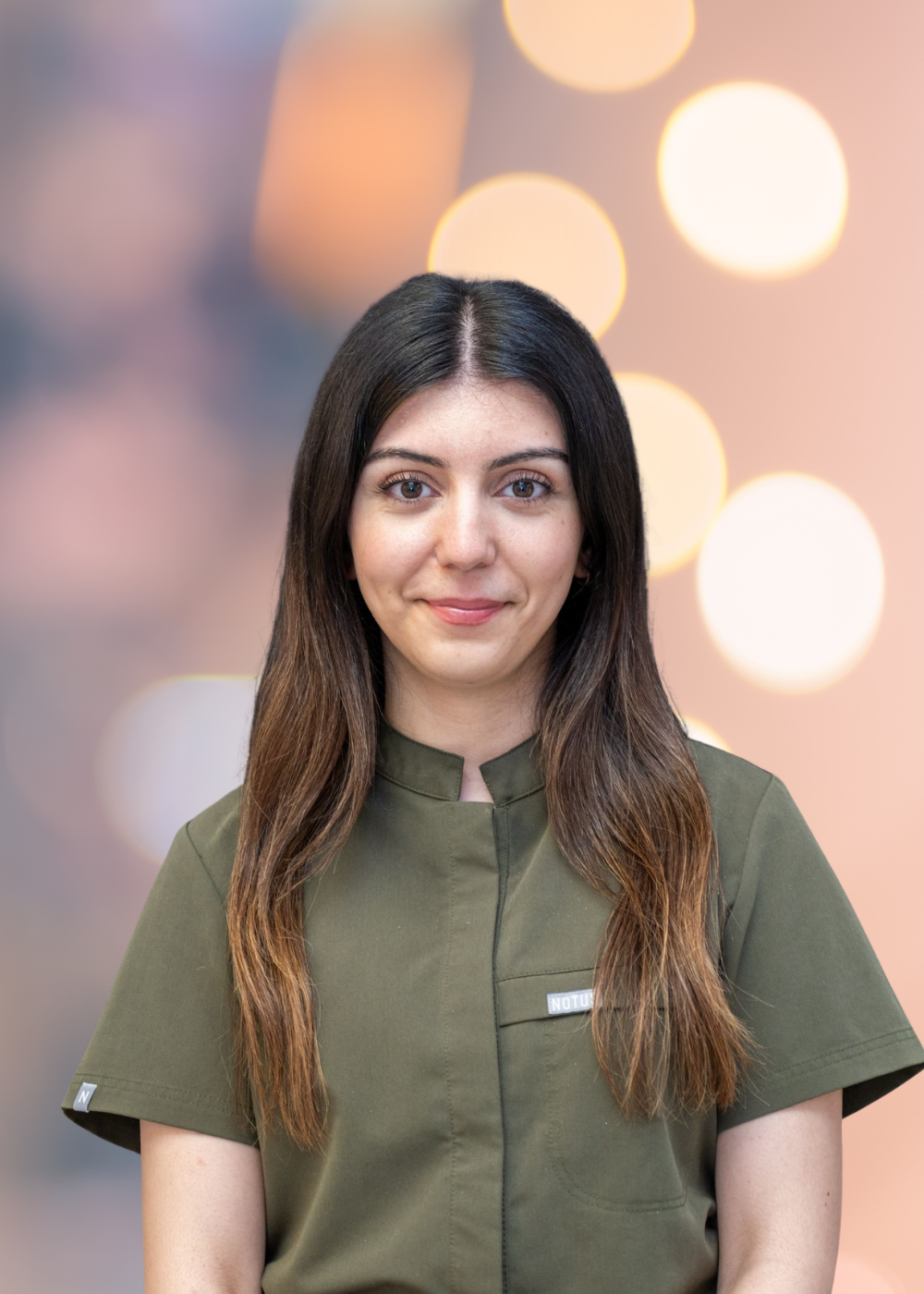 Portrait of a smiling woman with long dark hair wearing olive-green scrubs, facing the camera with a blurred warm background.
