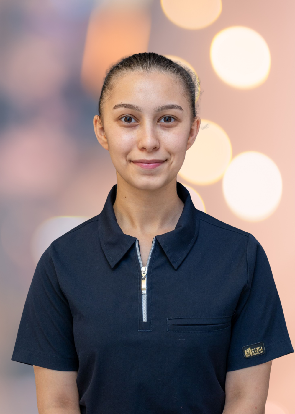 Portrait of a young woman with dark hair tied back, wearing a navy polo, against a soft bokeh background.