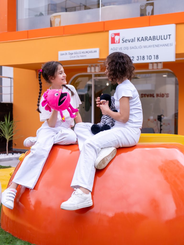 Two smiling girls in white outfits sit on an orange rounded structure outside a dental clinic, each holding a plush toy.